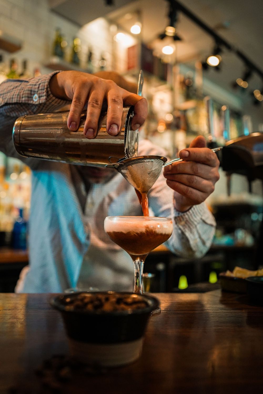 Bartender pours dark cocktail from shaker through strainer into glass at dimly lit bar counter.