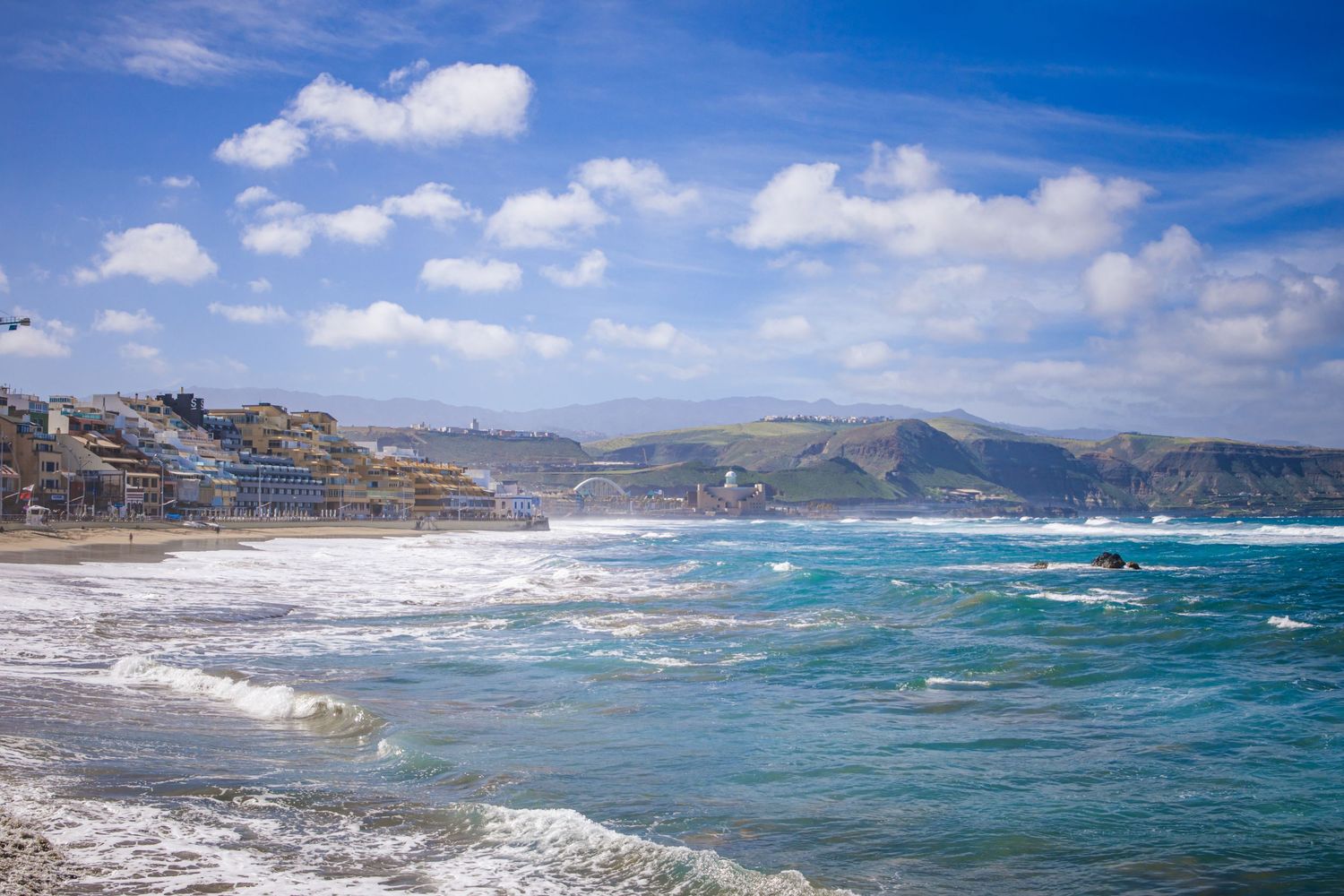 Turquoise waves crash against the rocky coastline of a Mediterranean seaside town under bright blue skies and white clouds.