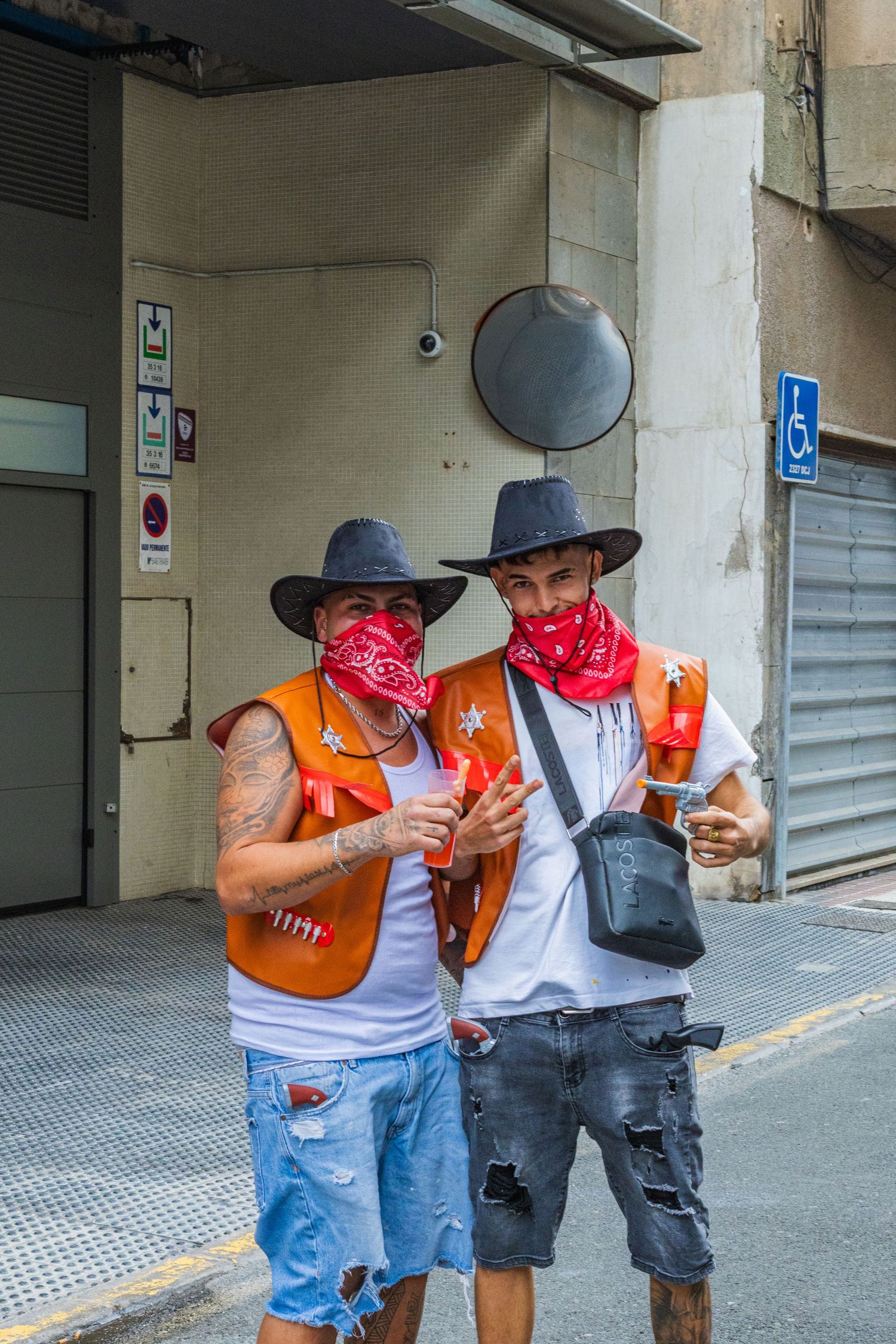 Two people in cowboy hats and casual attire share drinks while standing in an urban alleyway.