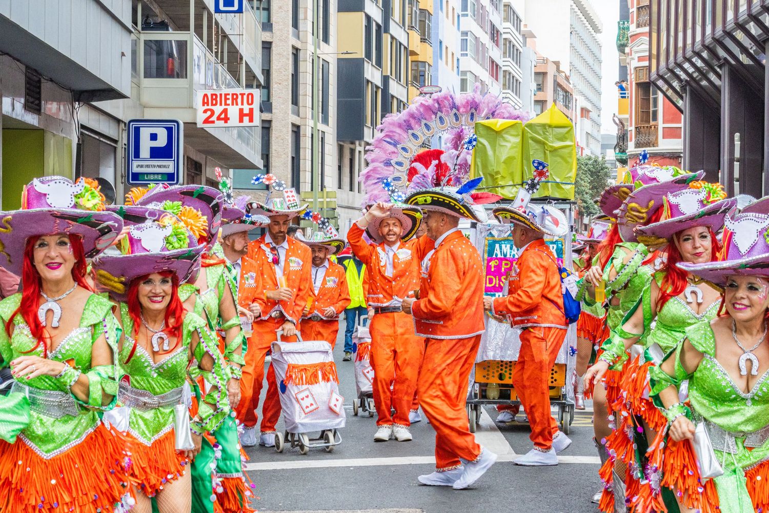 Colorful parade performers in bright orange and green costumes dance through city streets during a festival celebration.