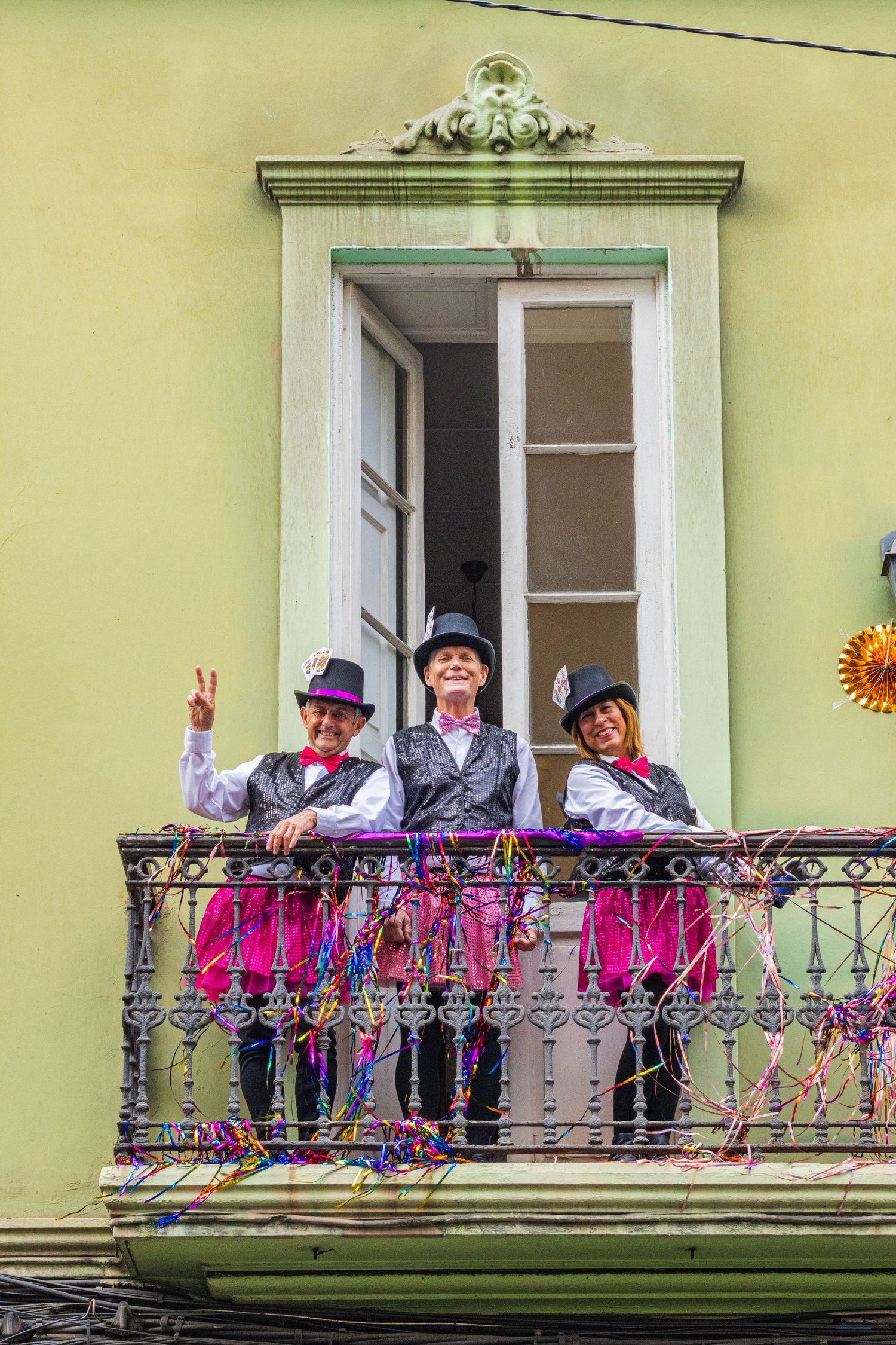 A group of people in colorful outfits and hats wave from a decorative balcony on a yellow building.