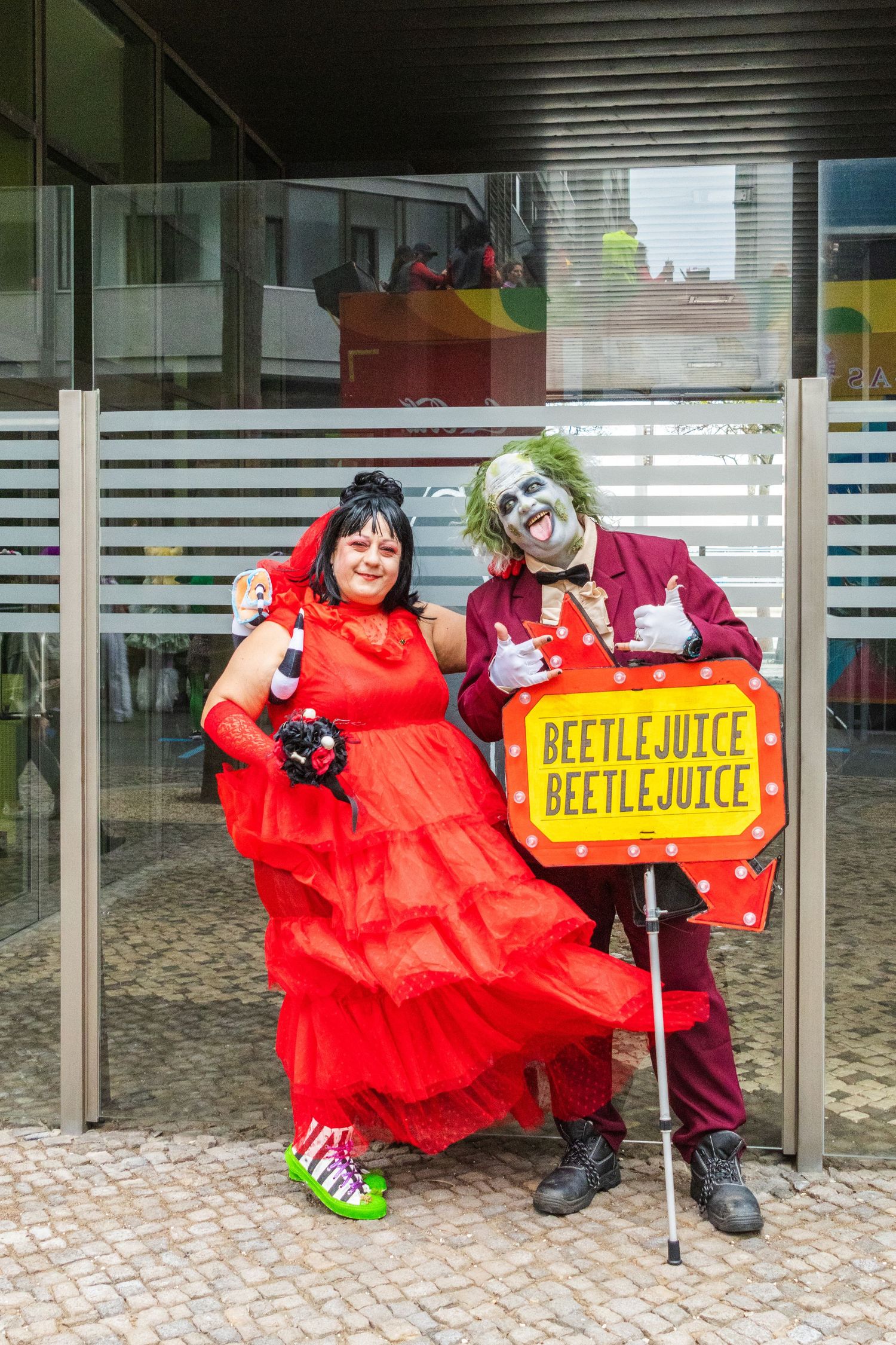 Two cosplayers dressed as characters pose next to a Beetlejuice sign at a convention.