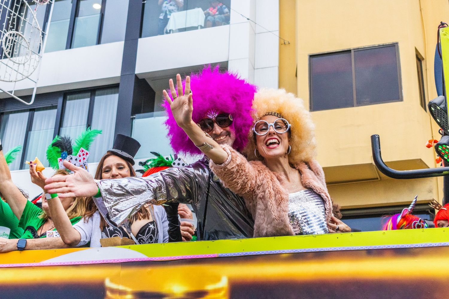 Colorfully dressed parade participants wave from a yellow float during a festive street celebration.