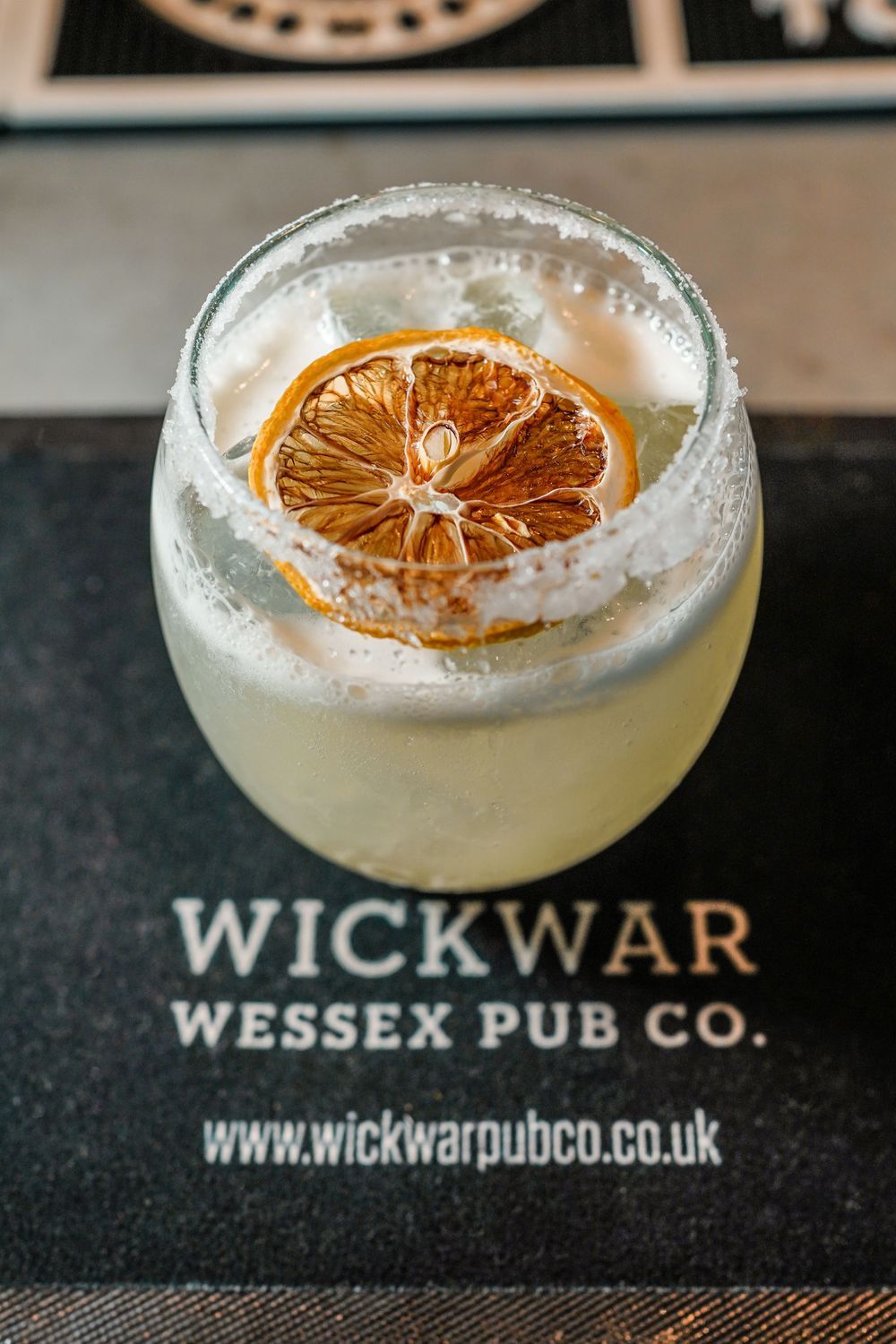 A craft cocktail with dried citrus garnish served on a Wickwar Wessex Pub Co. coaster with frothy white foam.