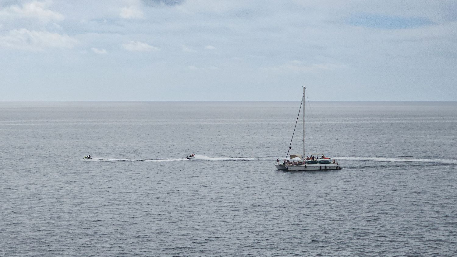 Sailboat navigates calm ocean waters under cloudy skies with distant boats visible on the horizon.