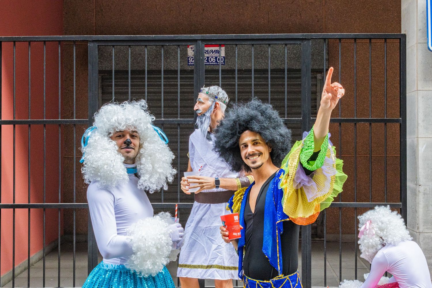 Three people in colorful costumes and silver wigs pose playfully in front of a metal gate.