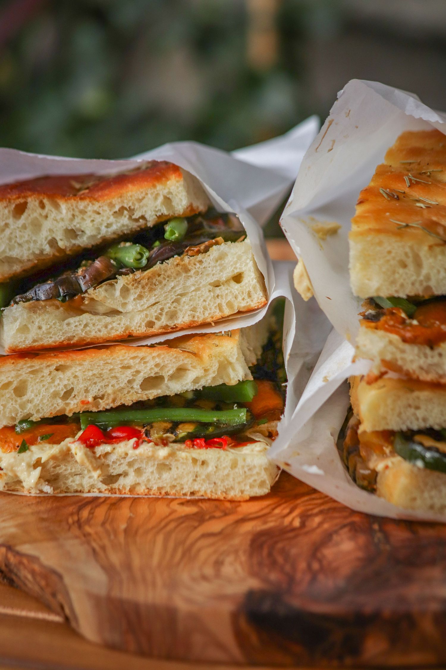 Fresh sandwich halves wrapped in paper on wooden board featuring colorful vegetables and fillings on golden bread.
