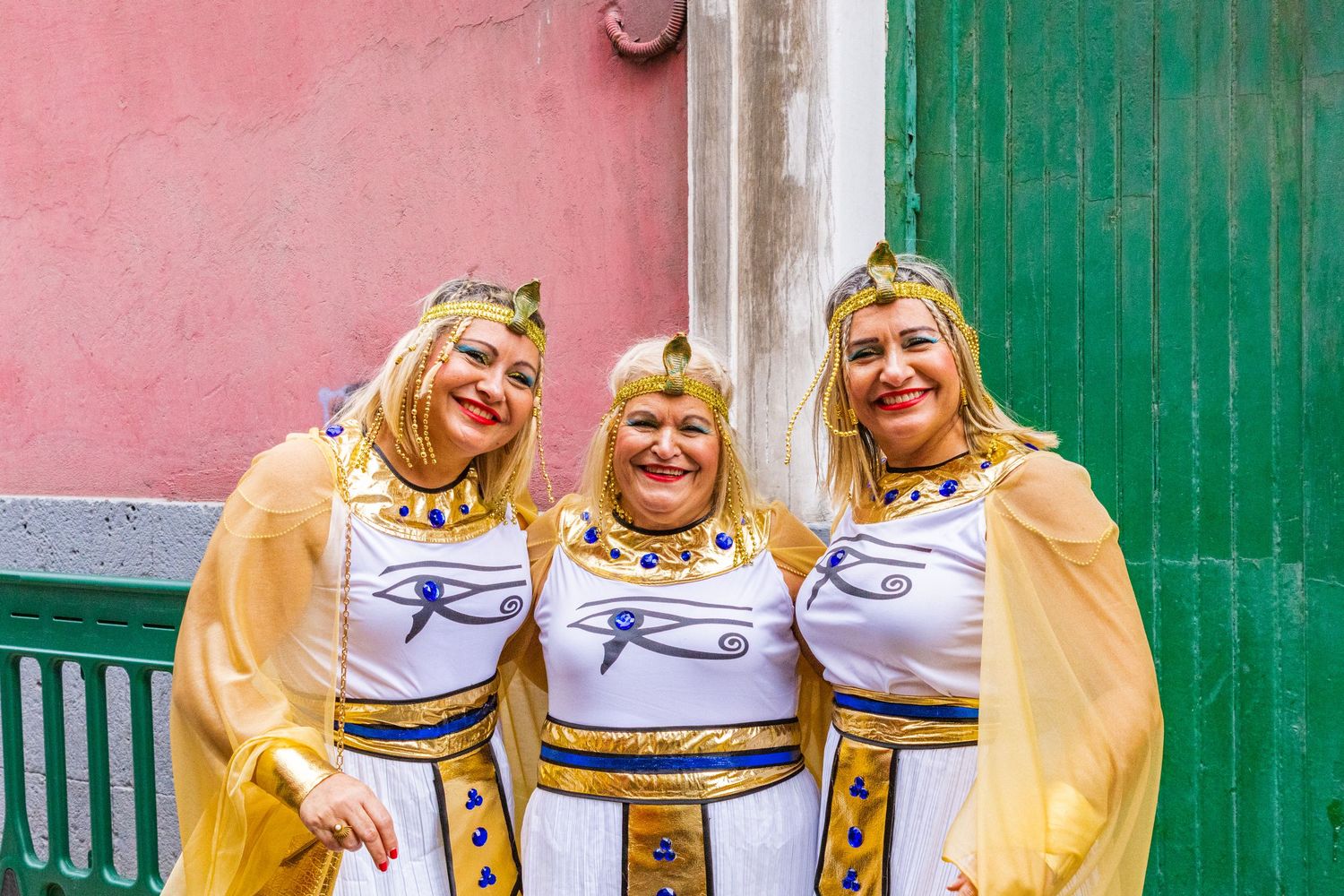 Three performers in matching yellow and white outfits smile and pose together against a pink and green wall.