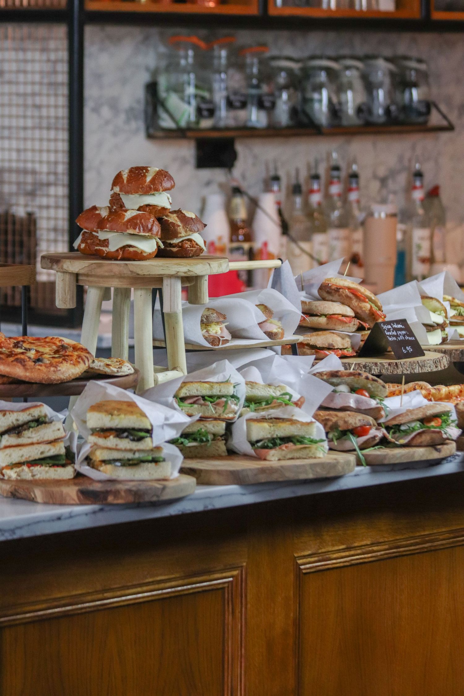 A restaurant counter displays various sandwiches and burgers on wooden boards with a bar shelf of liquor bottles in background.