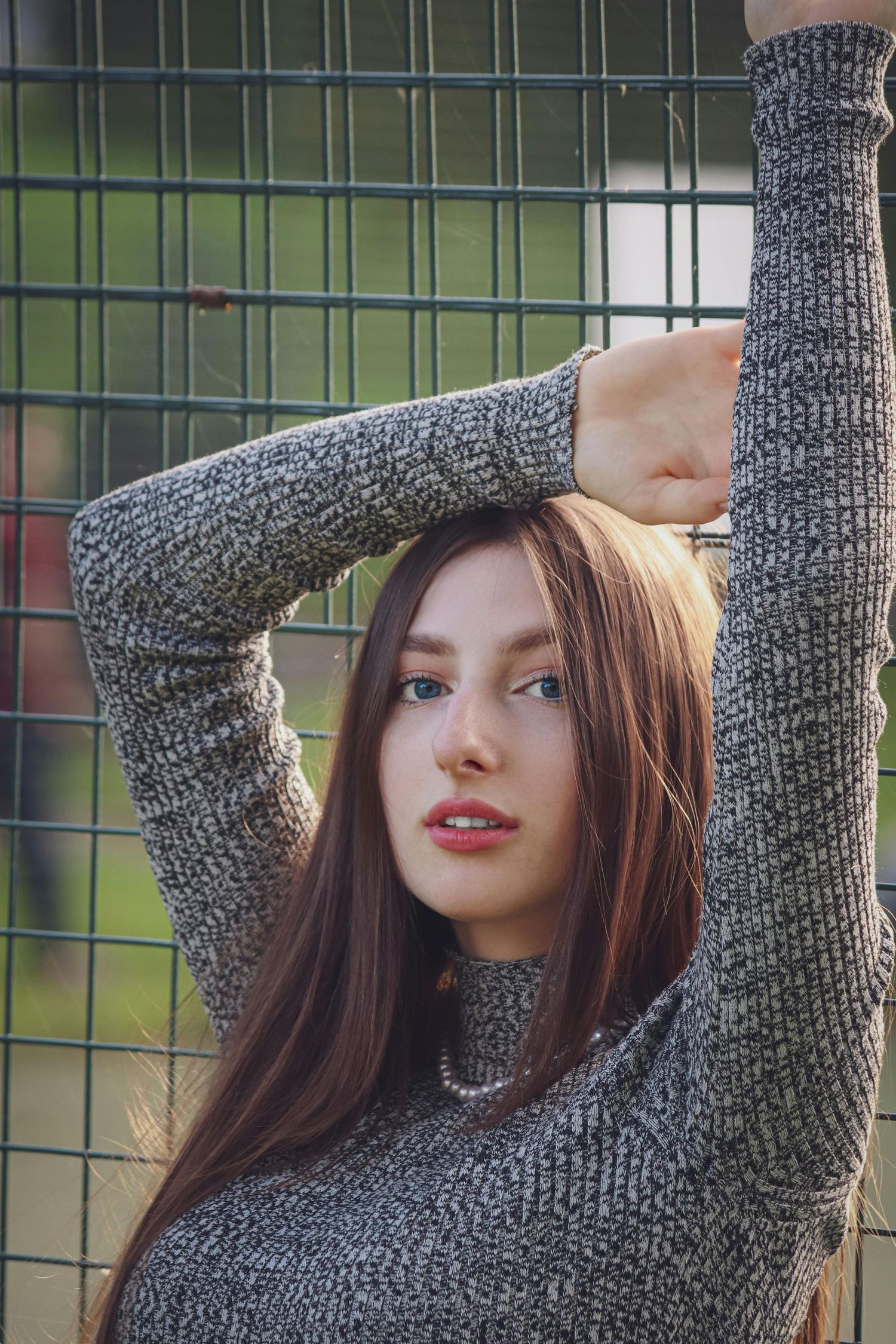 Person wearing a gray knit sweater poses against a fence with green foliage in the background.