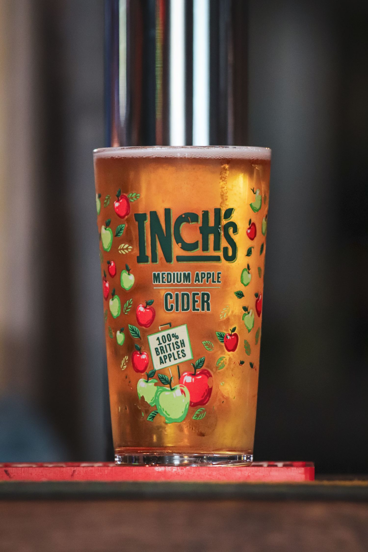 A glass of Inchs hard apple cider with colorful apple illustrations on the cup sits on a wooden surface in warm lighting.