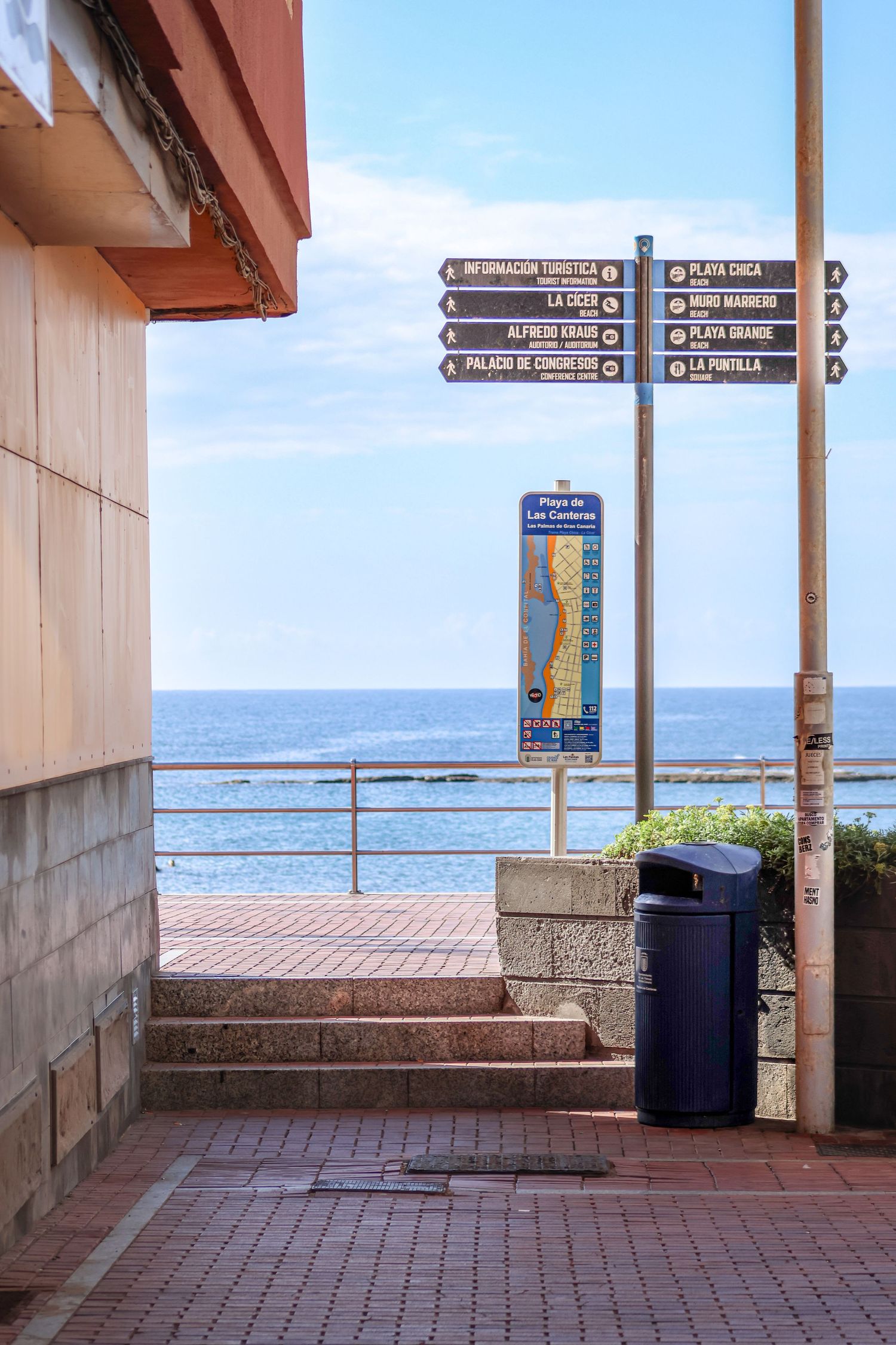 Covered walkway with brick pavers and directional signs overlooking the ocean and distant coastline.