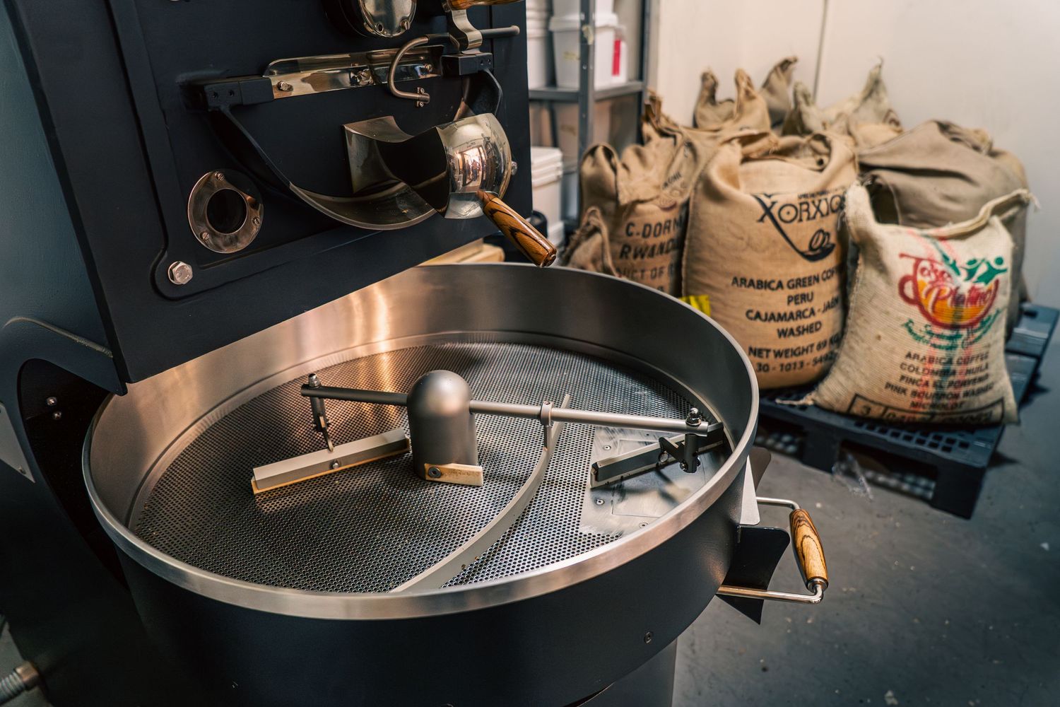 Coffee roaster machine with tools and burlap sacks of green coffee beans in a roastery workshop.