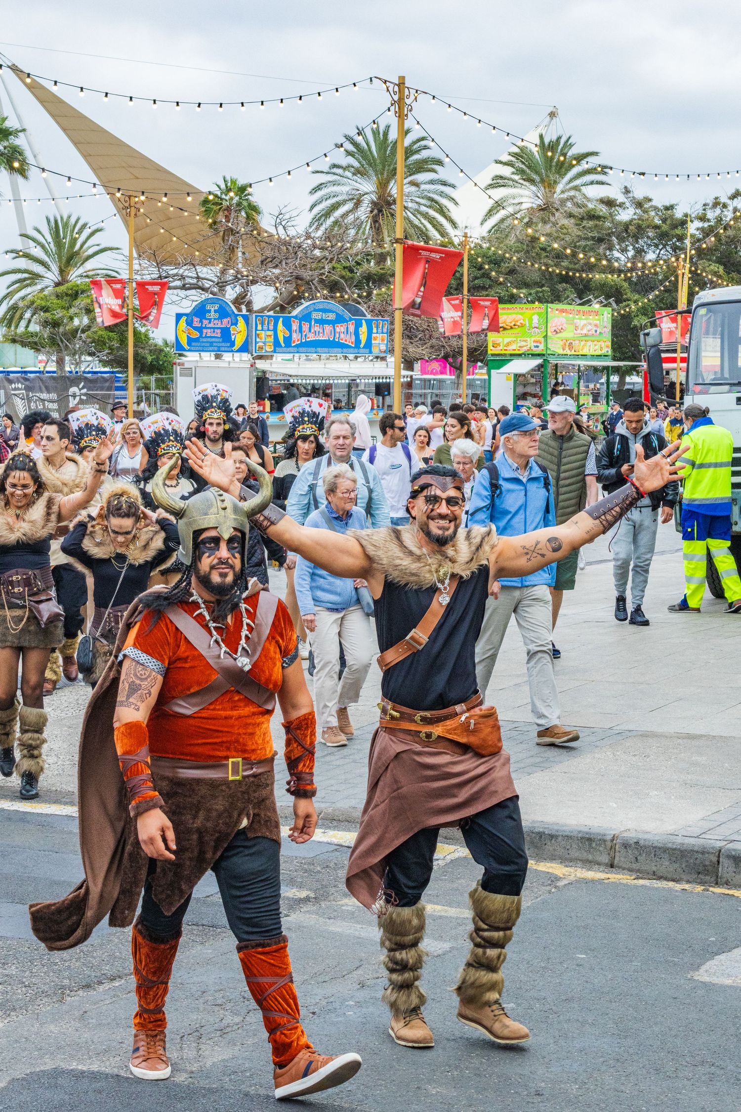 A group of costumed performers dance and entertain visitors at an outdoor festival with palm trees and string lights.