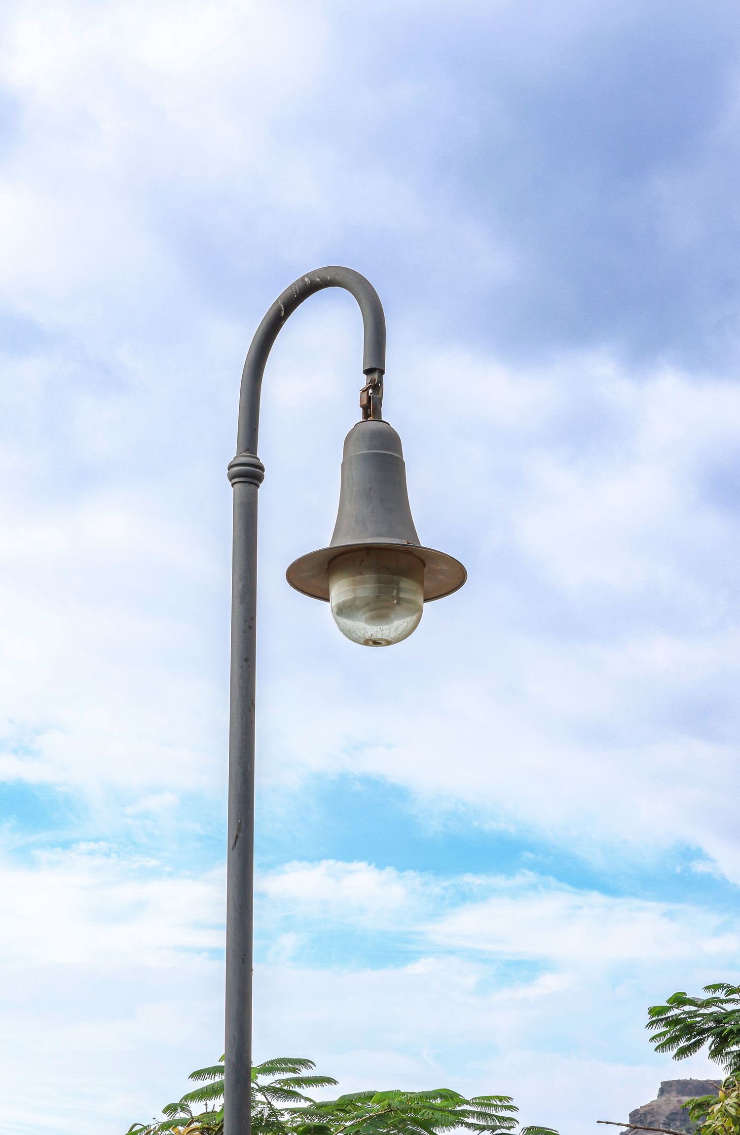 A vintage-style street lamp with curved pole stands against a blue cloudy sky with trees visible below.