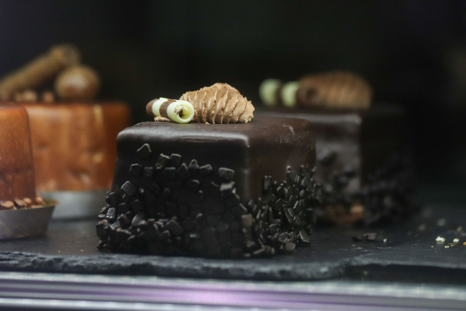 A dark chocolate dessert bar with black decorative details sits on a slate surface next to other confections.