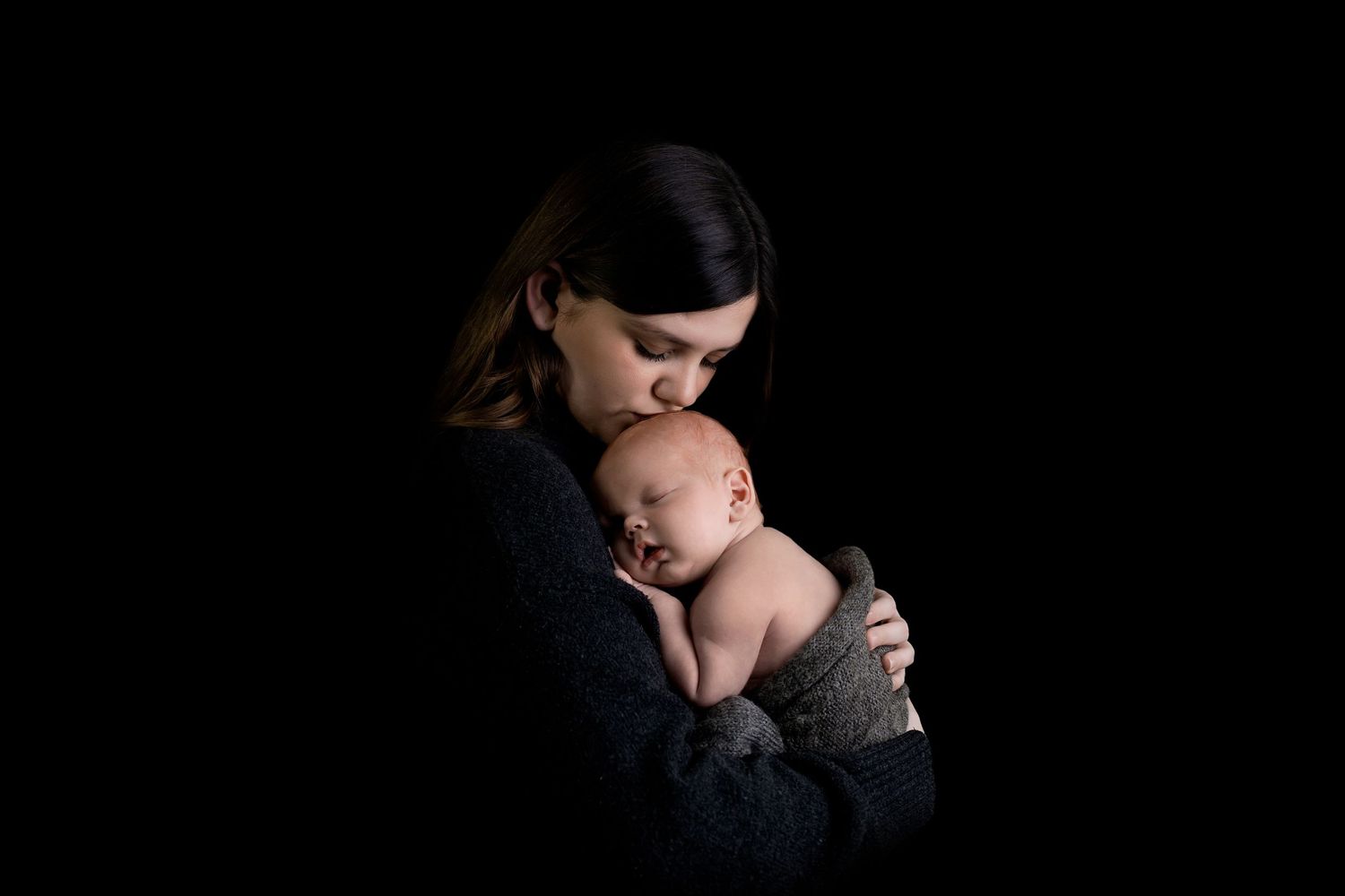 A tender moment captured in low-key lighting shows a parent gently embracing their sleeping newborn against a black background.
