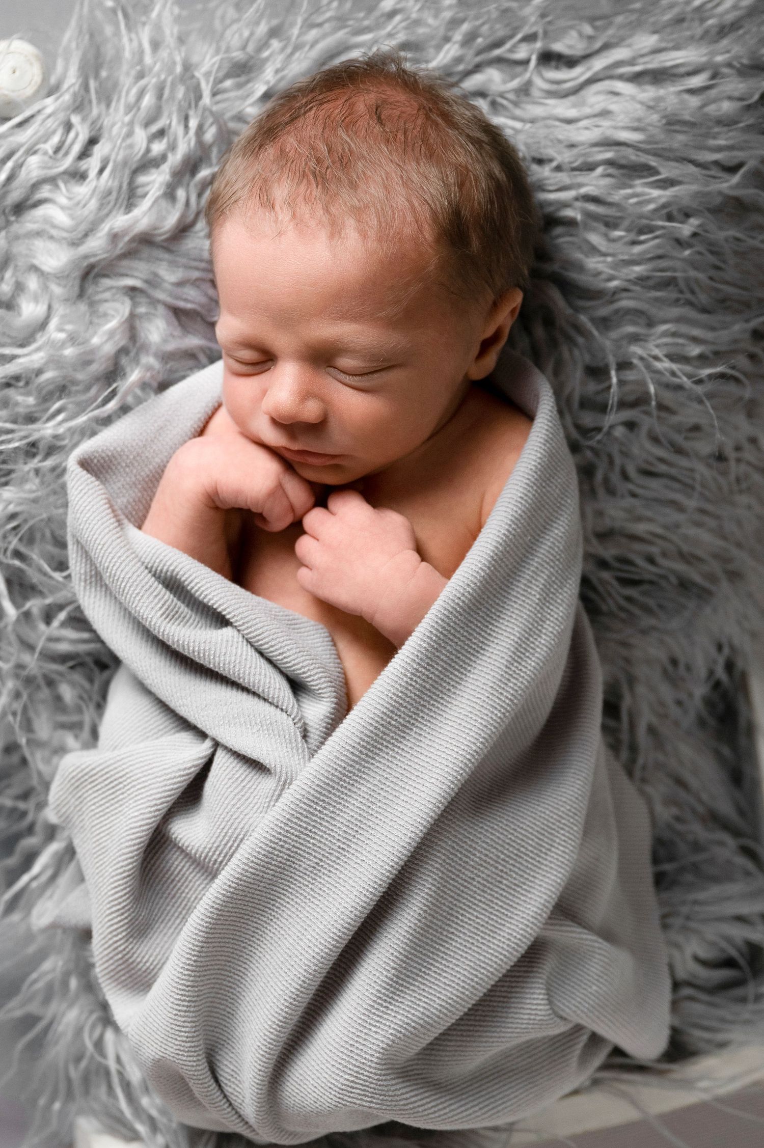 Peaceful newborn sleeping while swaddled in gray blanket on fluffy backdrop.