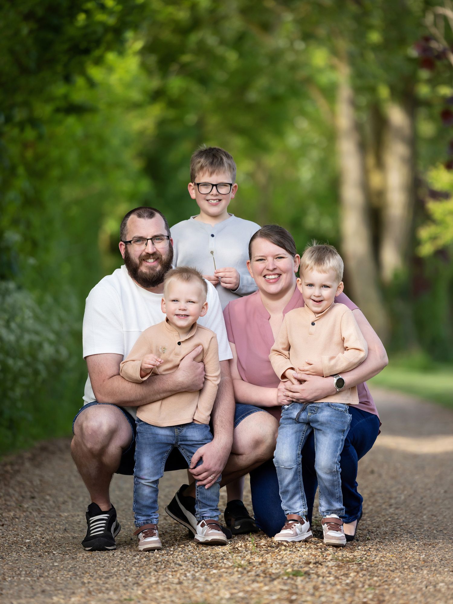 Happy family of five posing on a tree-lined path, parents with three young boys smiling outdoors in summer.