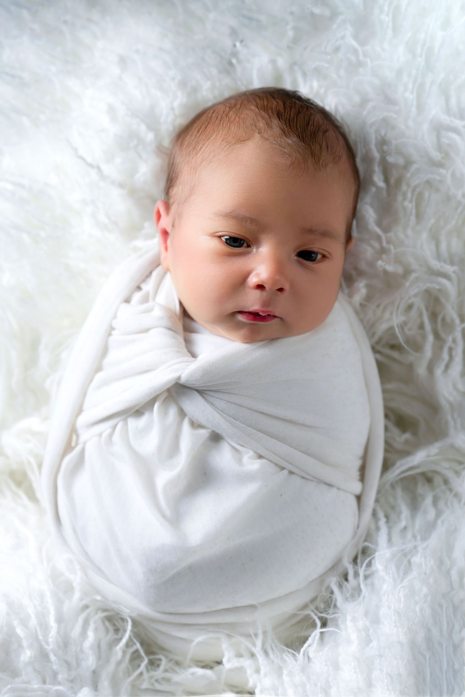 Newborn baby wrapped in white swaddle on fluffy white backdrop.