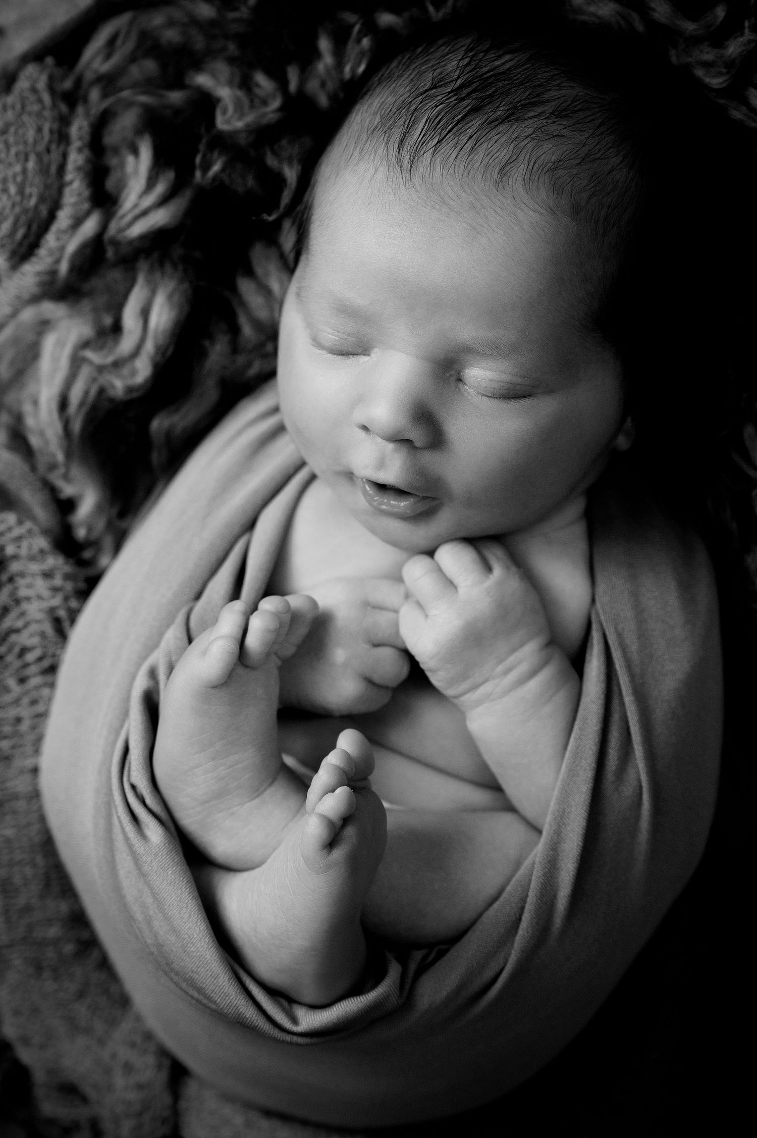 Artistic black and white photo of a peaceful newborn wrapped in fabric with tiny feet visible.