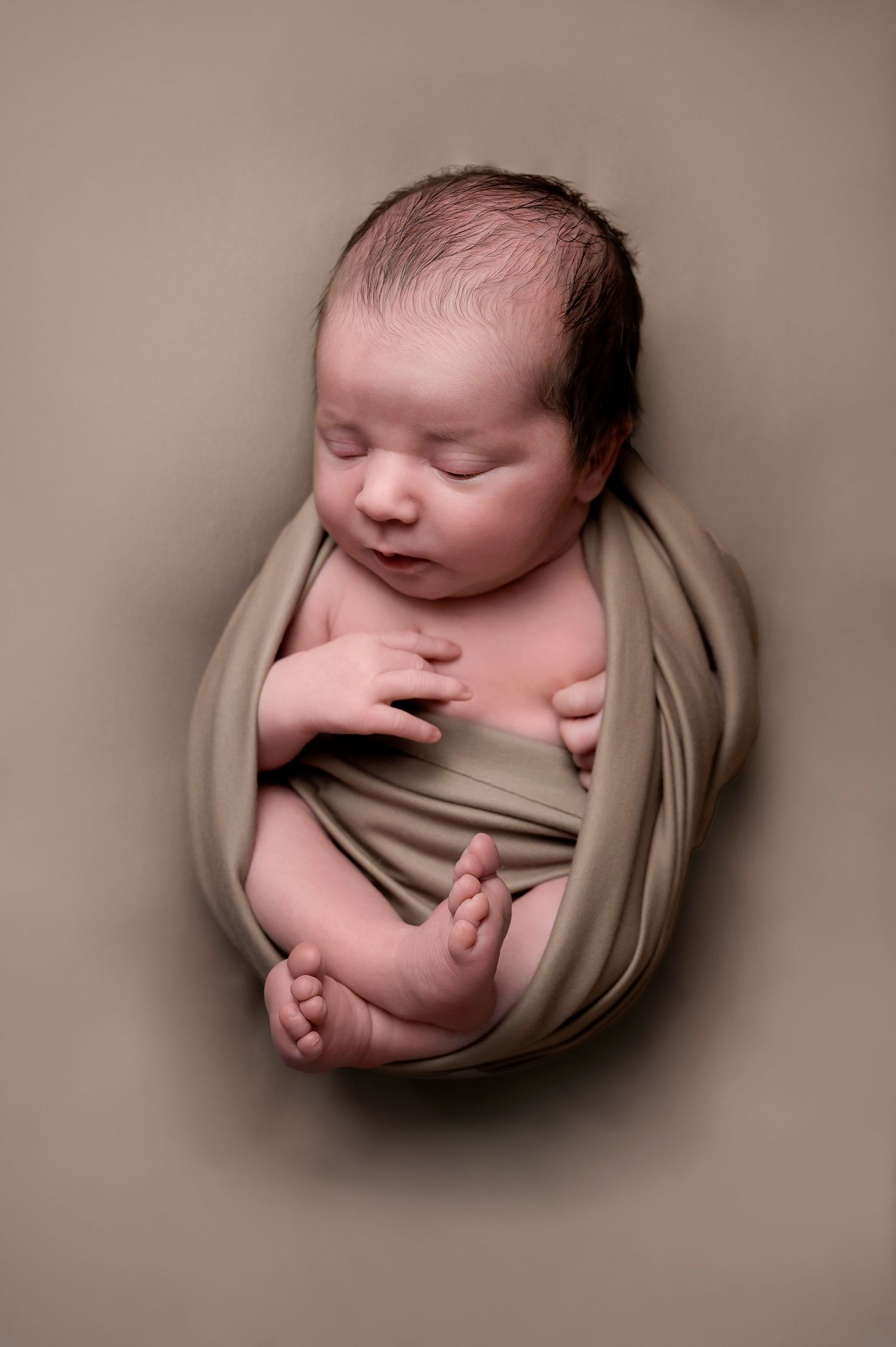 Sleeping newborn wrapped in beige fabric against neutral background in artistic pose.