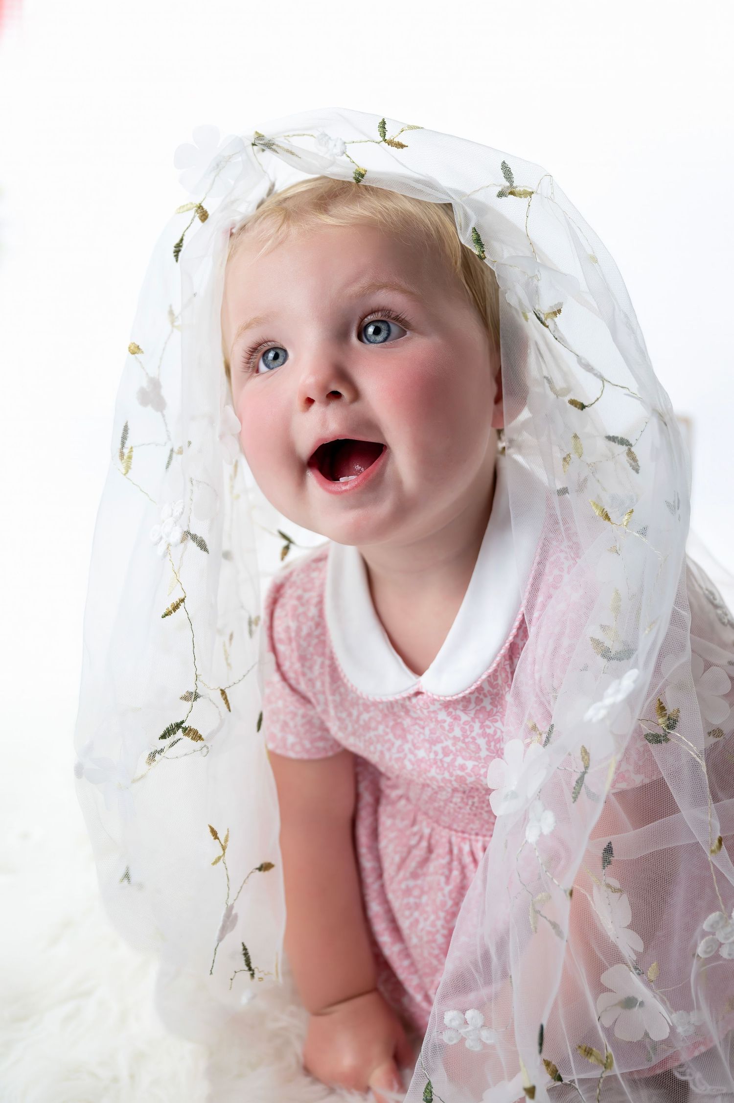 Joyful baby in pink dress peeks out from under delicate white floral veil.