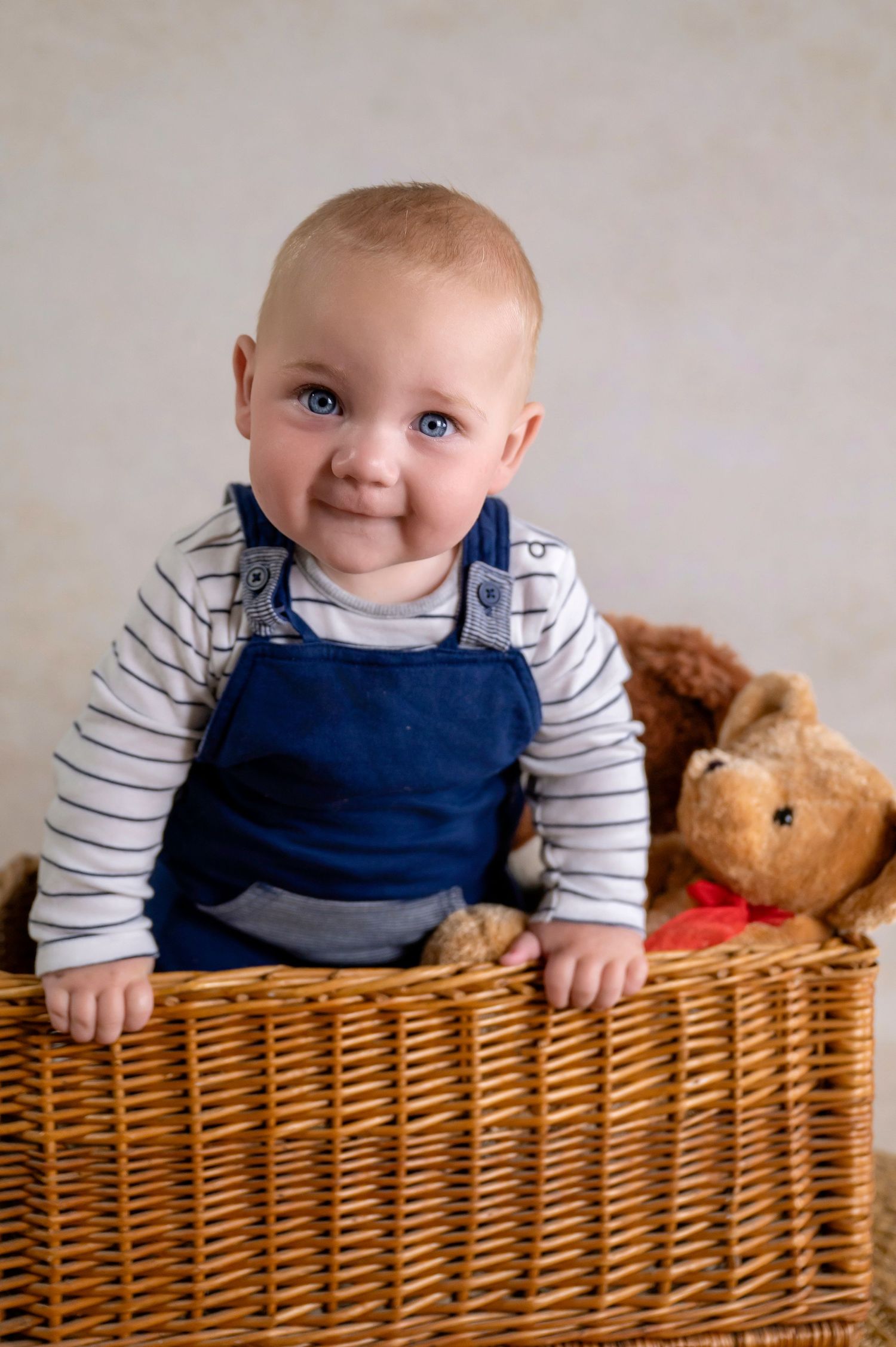 Smiling baby in blue overalls sitting in wicker basket with teddy bear.