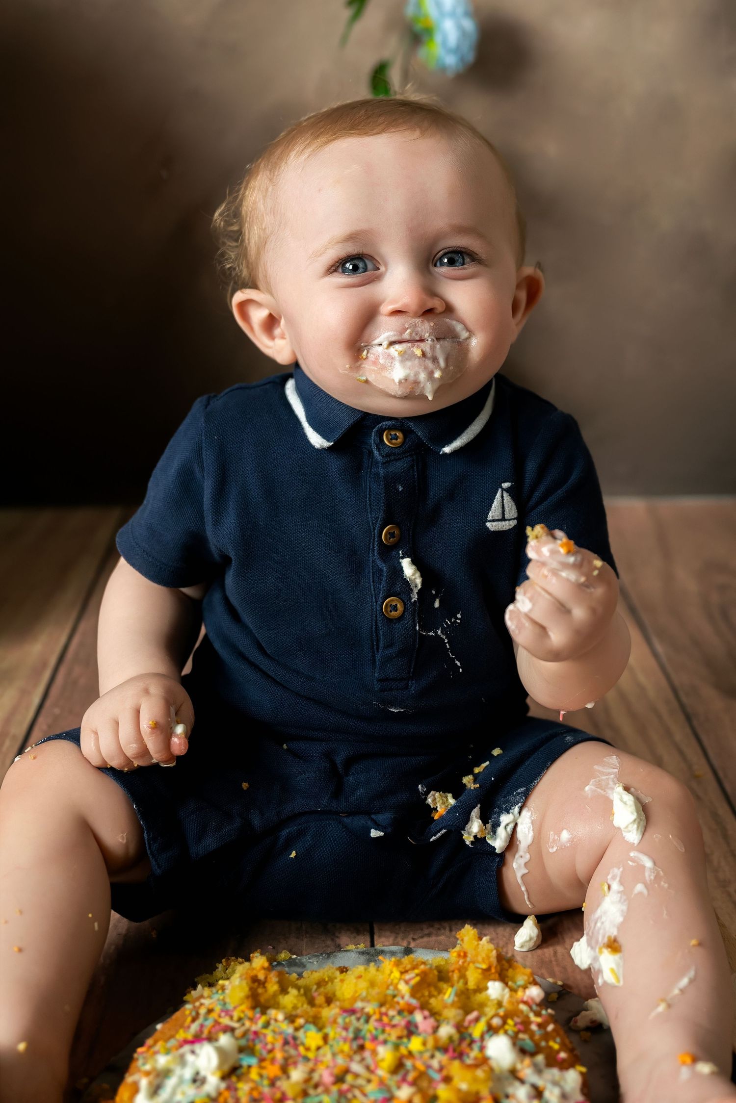 Messy baby in navy outfit with frosting on face sitting by destroyed cake with sprinkles.