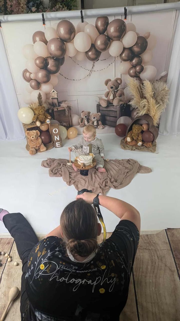 Photographer capturing a baby's first birthday cake smash with teddy bear theme, balloons, and ONE sign backdrop.