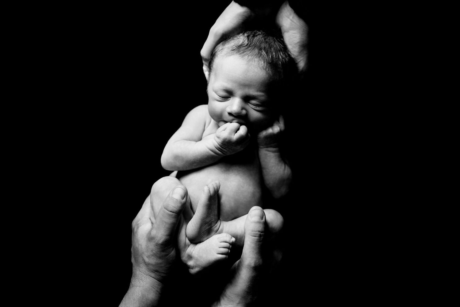 Artistic black and white photo of tiny newborn being gently held in dramatic lighting.