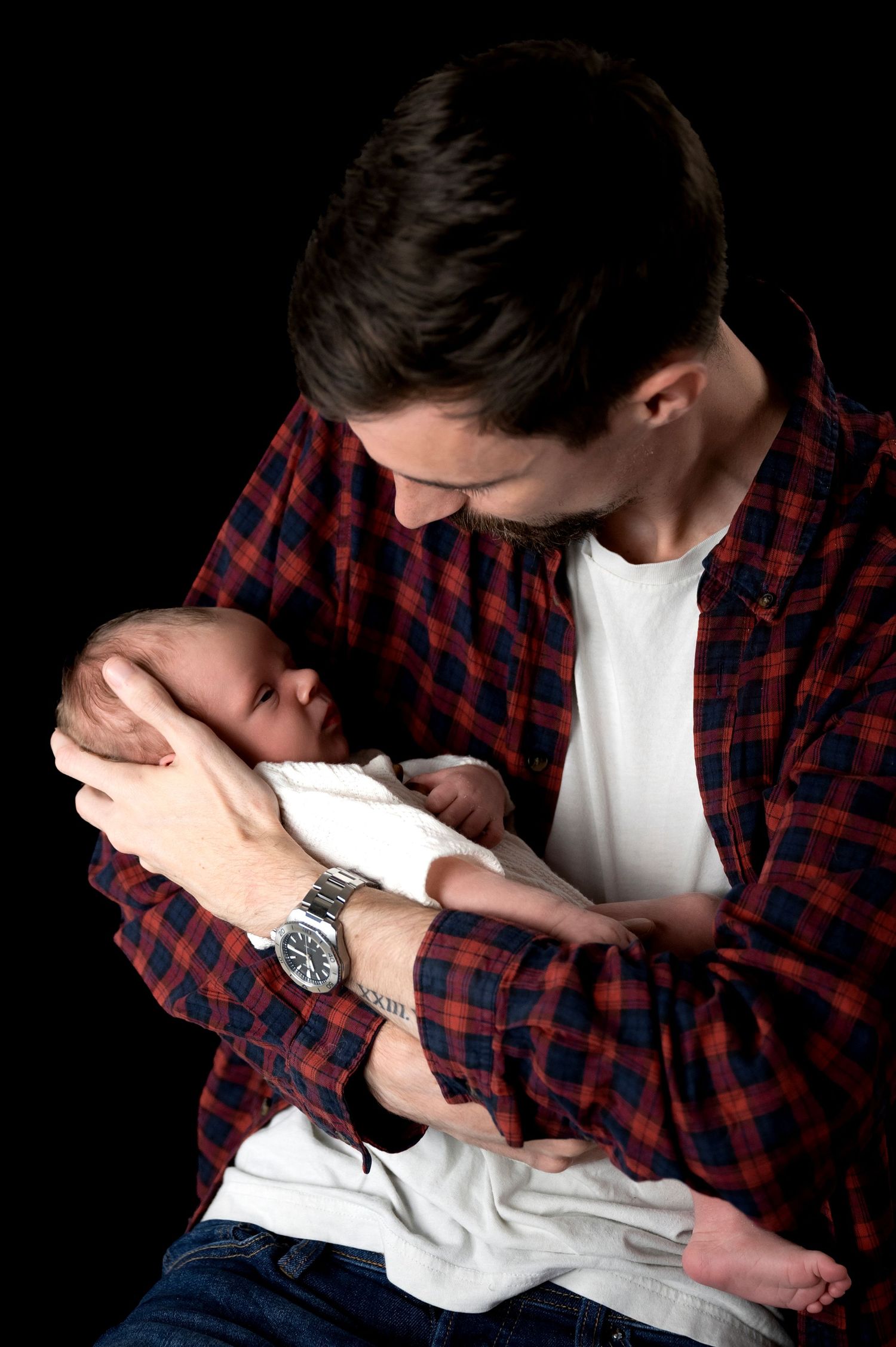 Tender moment between parent in plaid shirt holding newborn against black background.