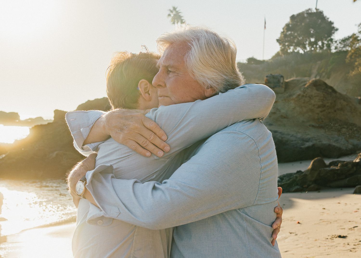 Two people embrace in a warm hug on a beach at sunset with rocks and trees visible in the background.