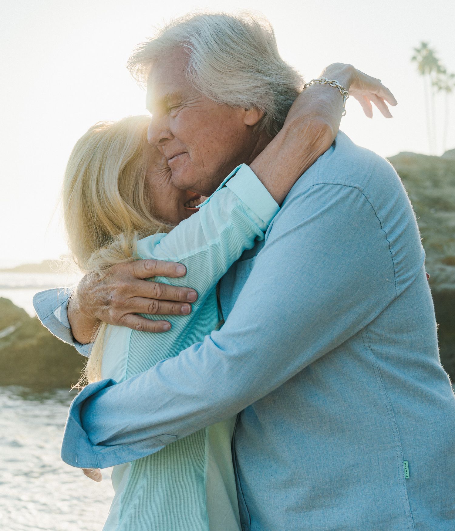 A couple embraces warmly on a beach at sunset, wearing light blue and turquoise sweaters.