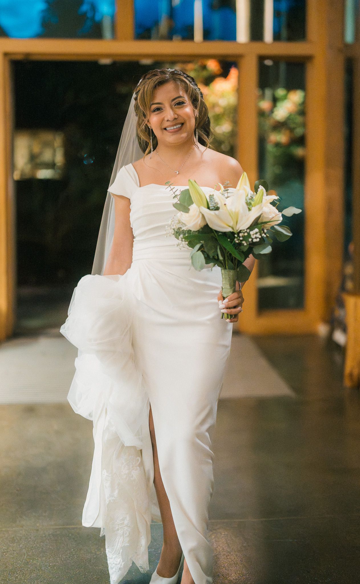 A bride in a white wedding dress holding a bouquet of white flowers in an elegant indoor venue.