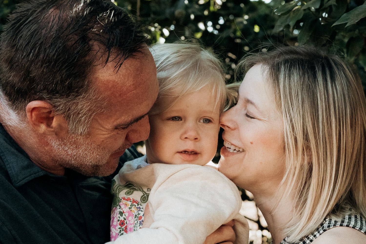 A family shares a tender moment together outdoors with their young child among green foliage.