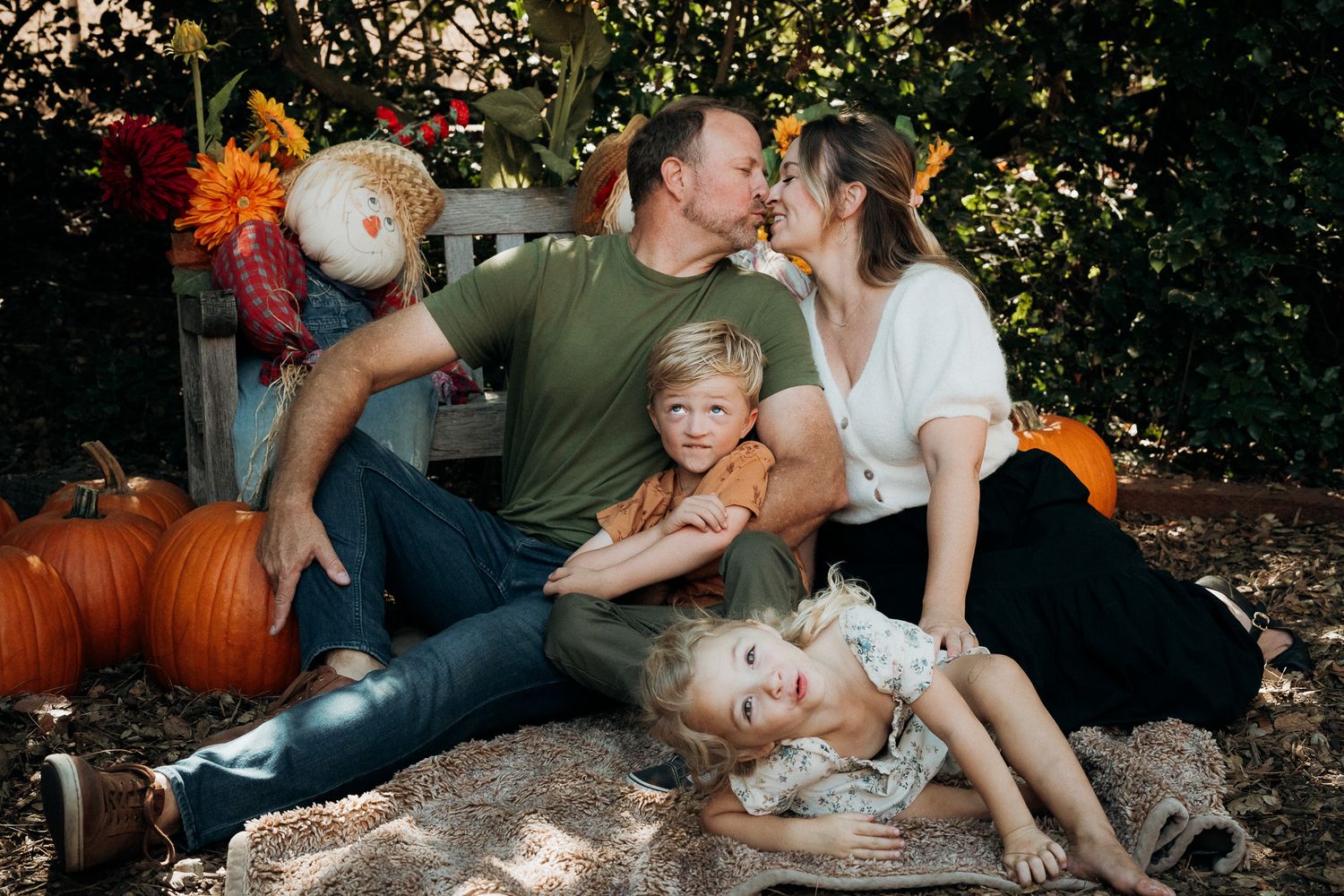 A family enjoys a fall photoshoot outdoors with pumpkins and autumn foliage in the background.