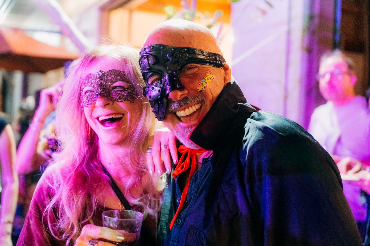 Two people in Halloween costumes including pink hair and a skull mask celebrate at a party with colorful lighting.