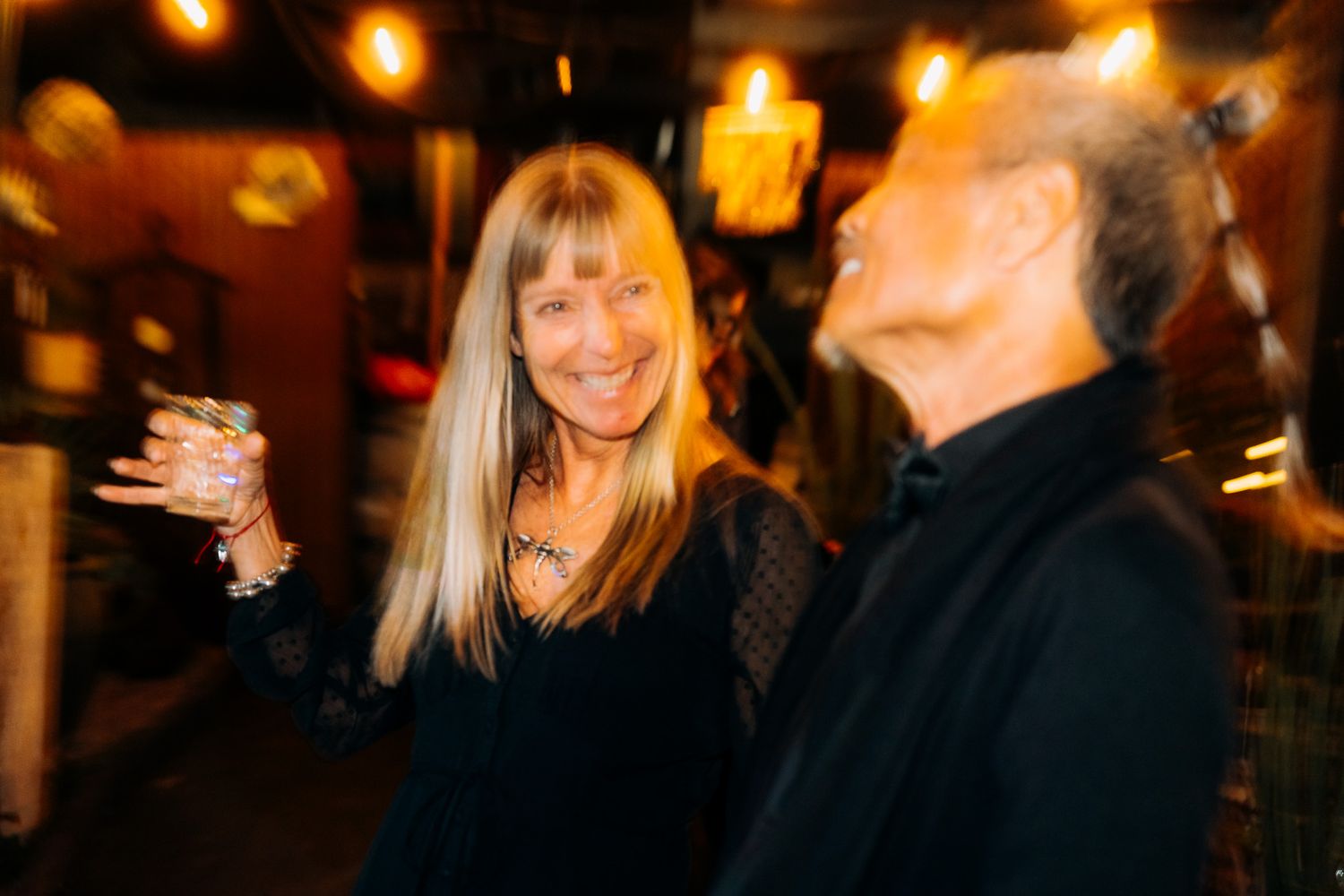 Two people laughing and socializing at a dimly lit bar or restaurant with warm ambient lighting in the background.