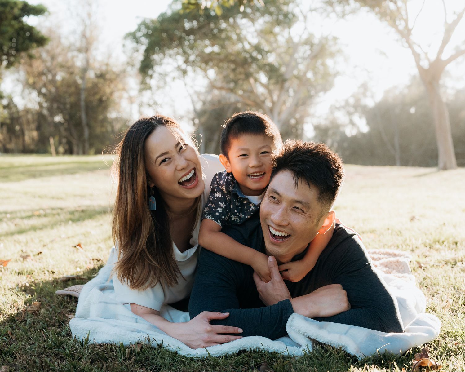 A family laughs together while laying on a white blanket in a sunlit park during a casual outdoor photo session.