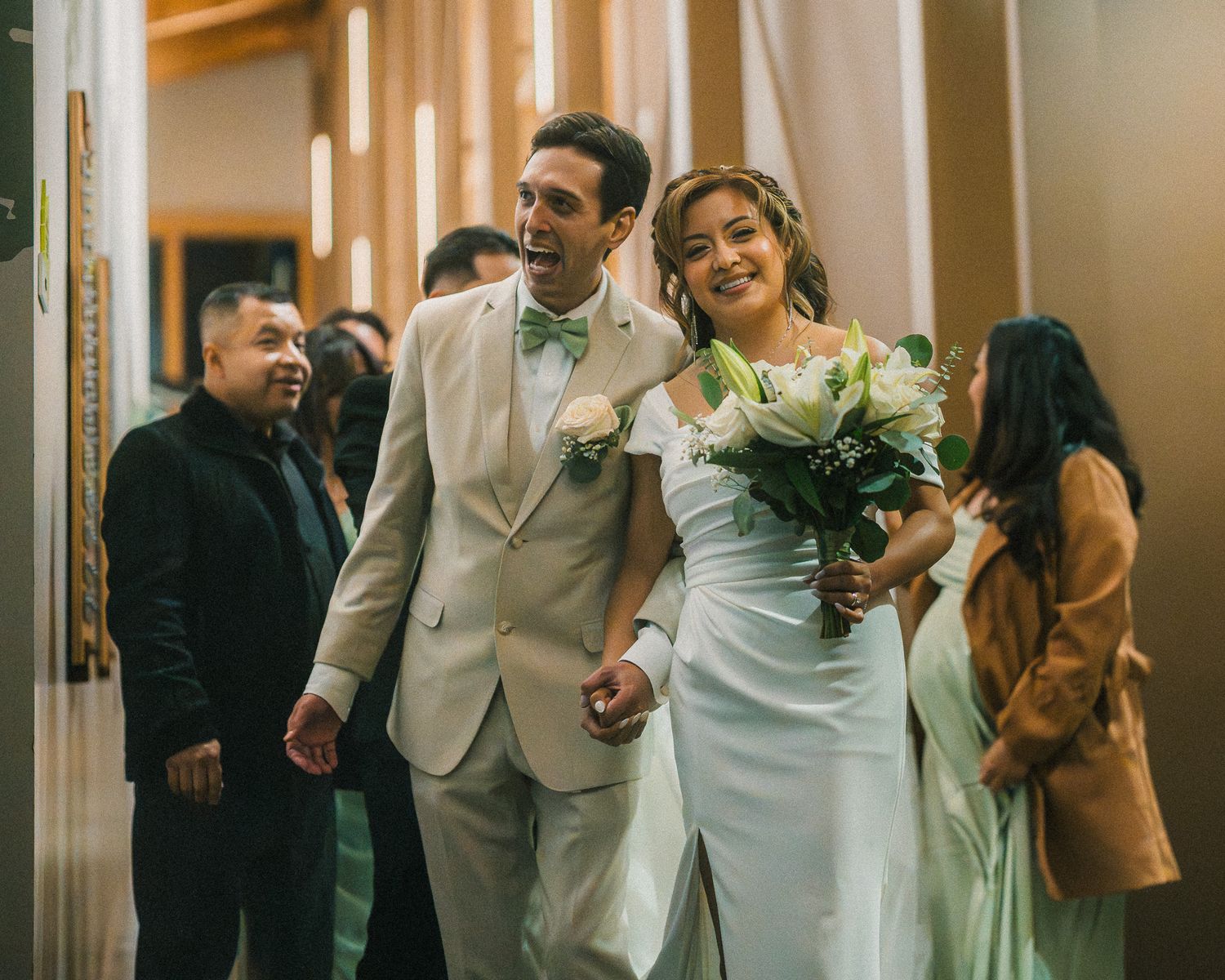 Wedding party walks down a hallway in formal attire with bridesmaids in sage green dresses and groomsmen in gray suits.