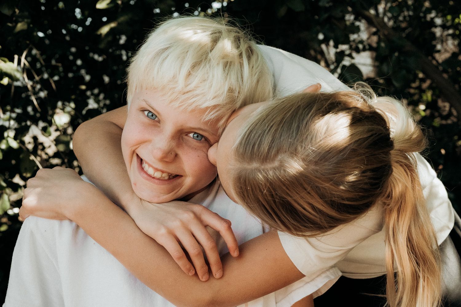 Two children share a joyful embrace outdoors with natural lighting and foliage in the background.