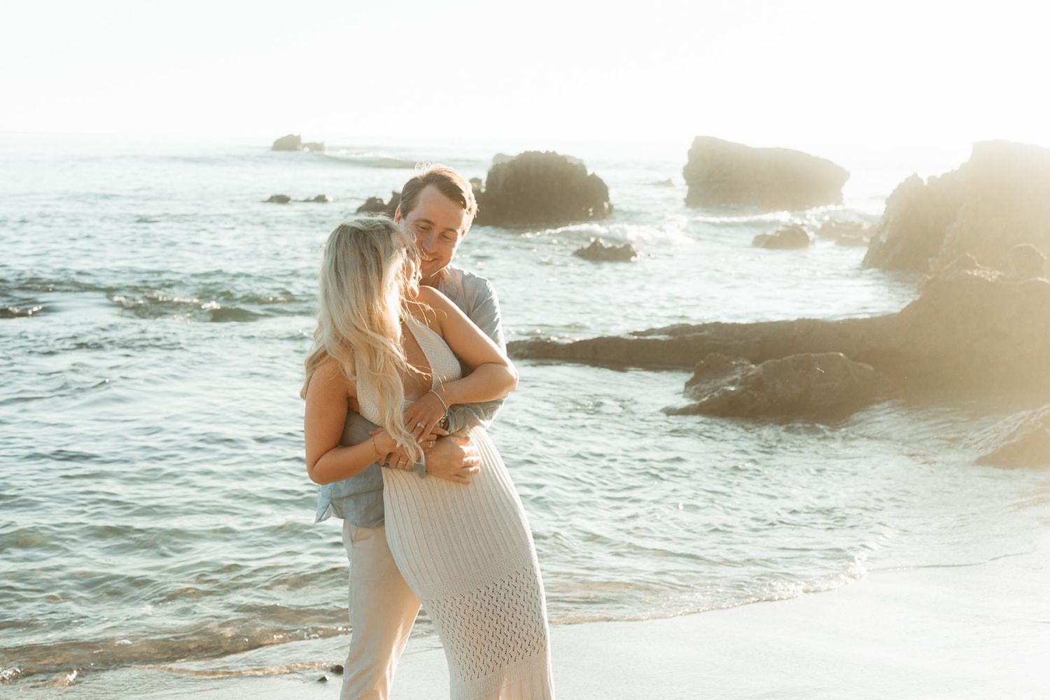 Romantic couple embracing on a scenic beach with rocky coastline and golden sunlight.