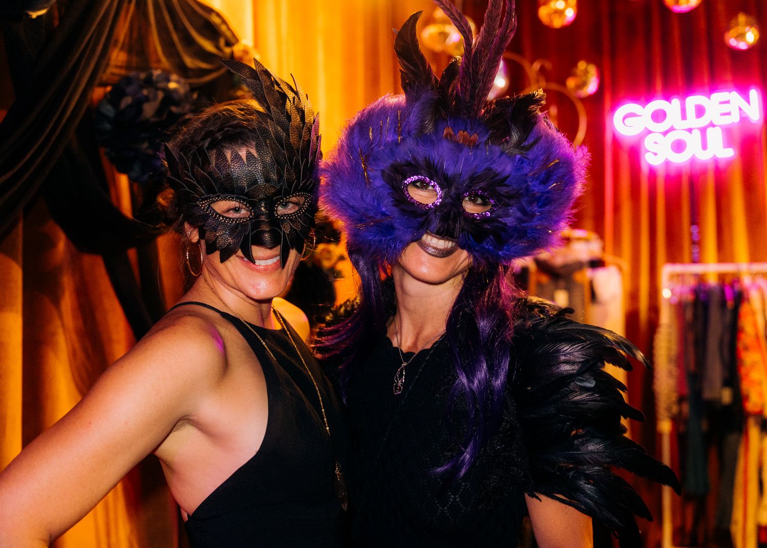 Masquerade party guests in black and purple feathered masks dance under warm orange lighting at an elegant event.