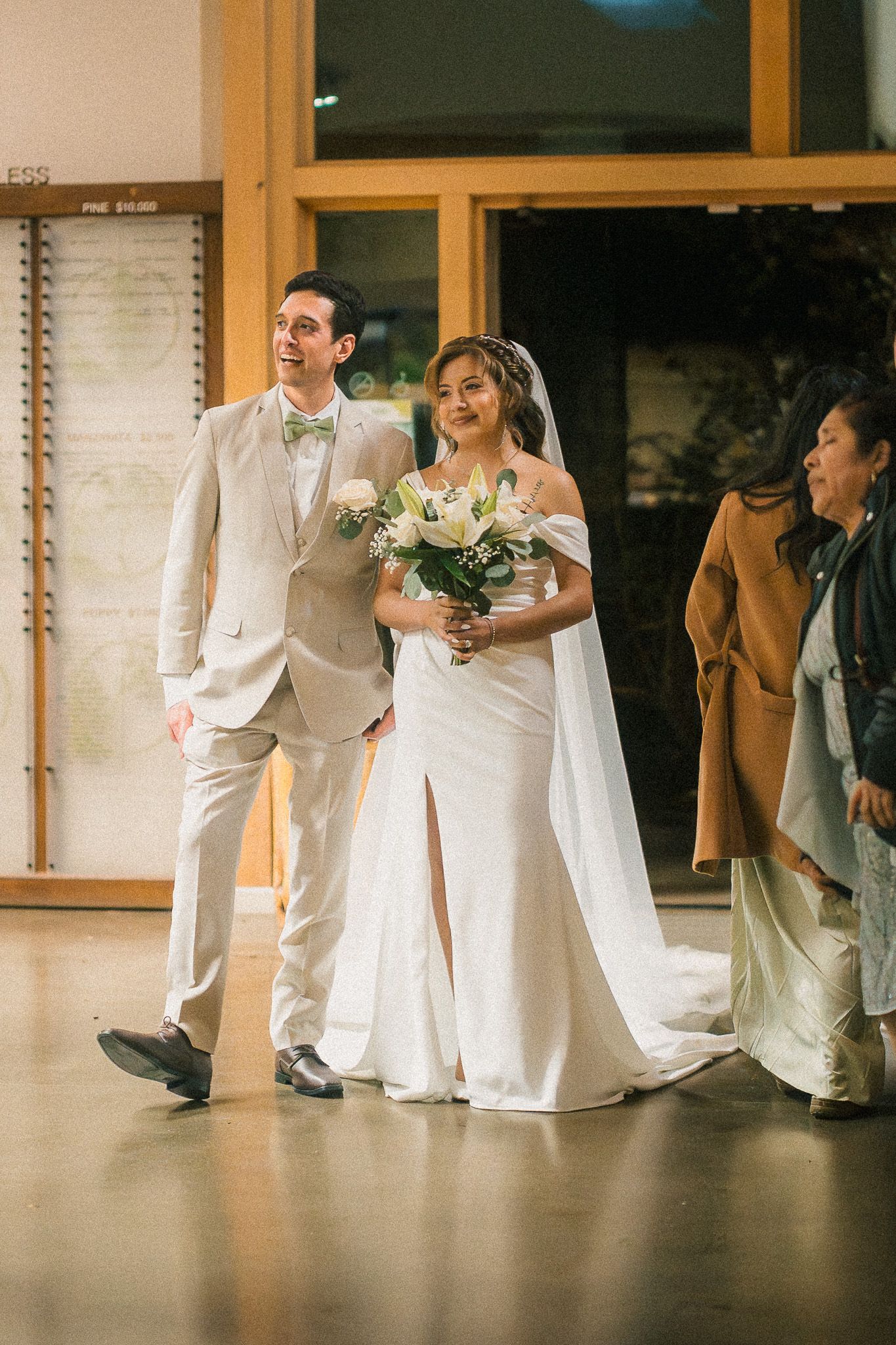 A couple walks together at their wedding ceremony, with the bride in a white gown holding a bouquet and the groom in a tan suit.