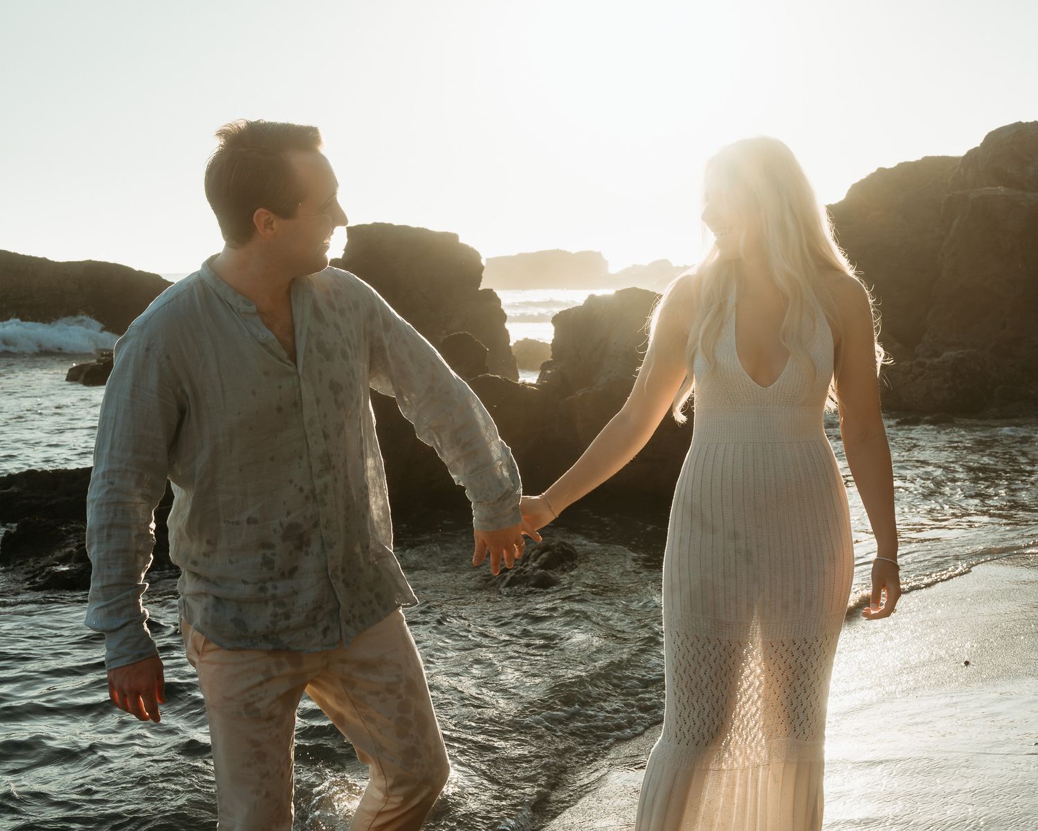 A romantic couple splashes in ocean waves at sunset with rocky cliffs in the background.