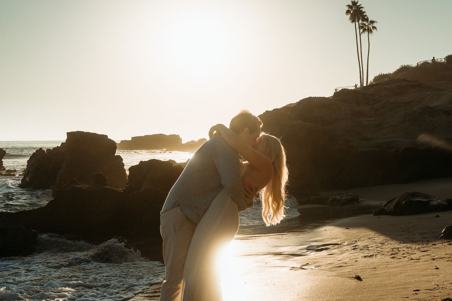 A romantic silhouette against the setting sun on a rocky beach with palm trees and ocean waves in the background.
