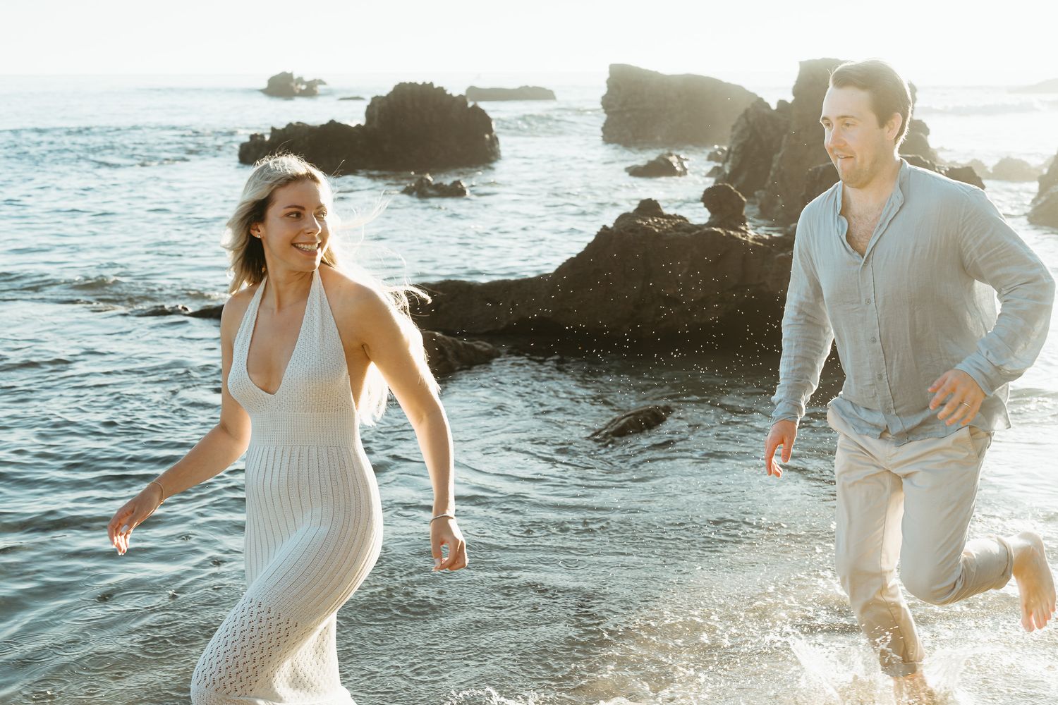 Couple running playfully along a rocky beach at sunset with waves and rock formations in the background.
