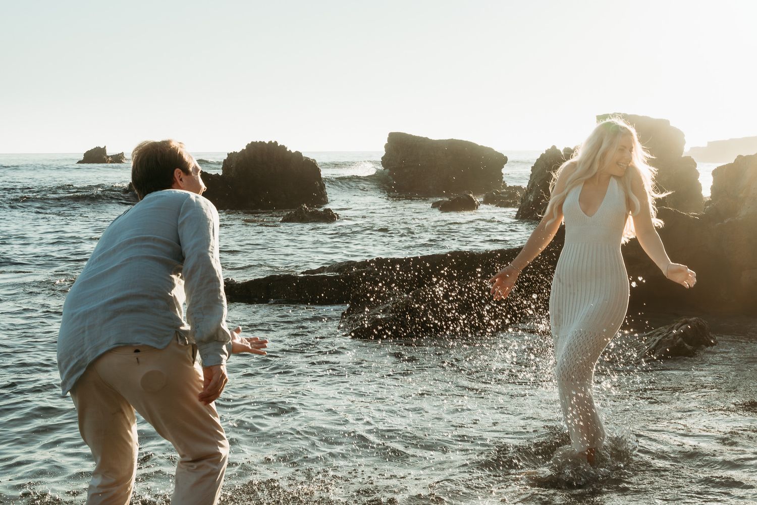 Couple playfully splashing in ocean waves among rocky coastline during golden hour.
