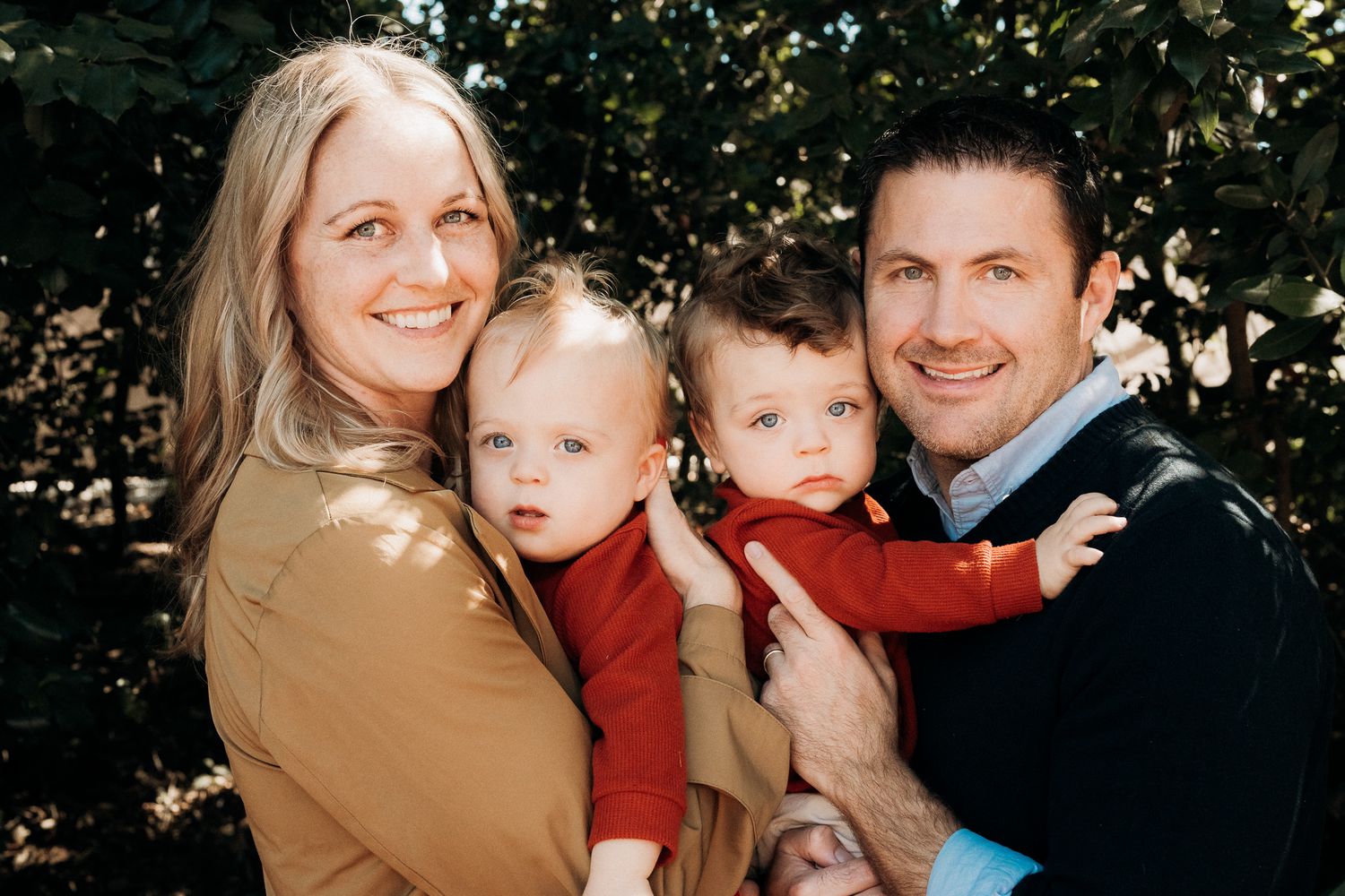 A family wearing coordinated tan and burgundy outfits poses together outdoors among trees.